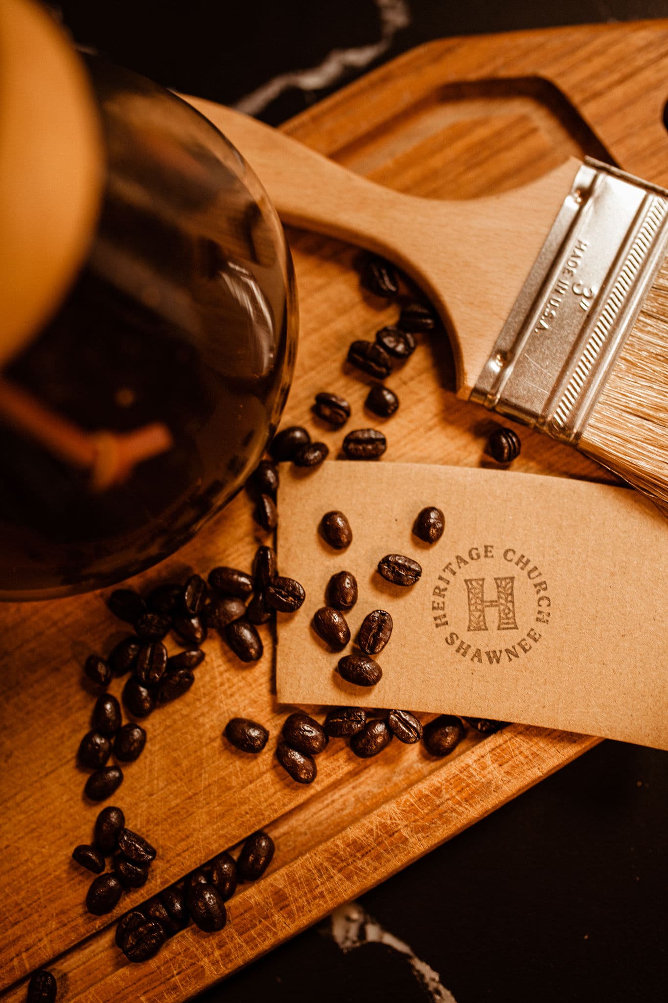 Dark roast whole coffee beans scattered around a Heritage Church Shawnee card on a wooden tray, evoking a cozy, artisanal coffee vibe.