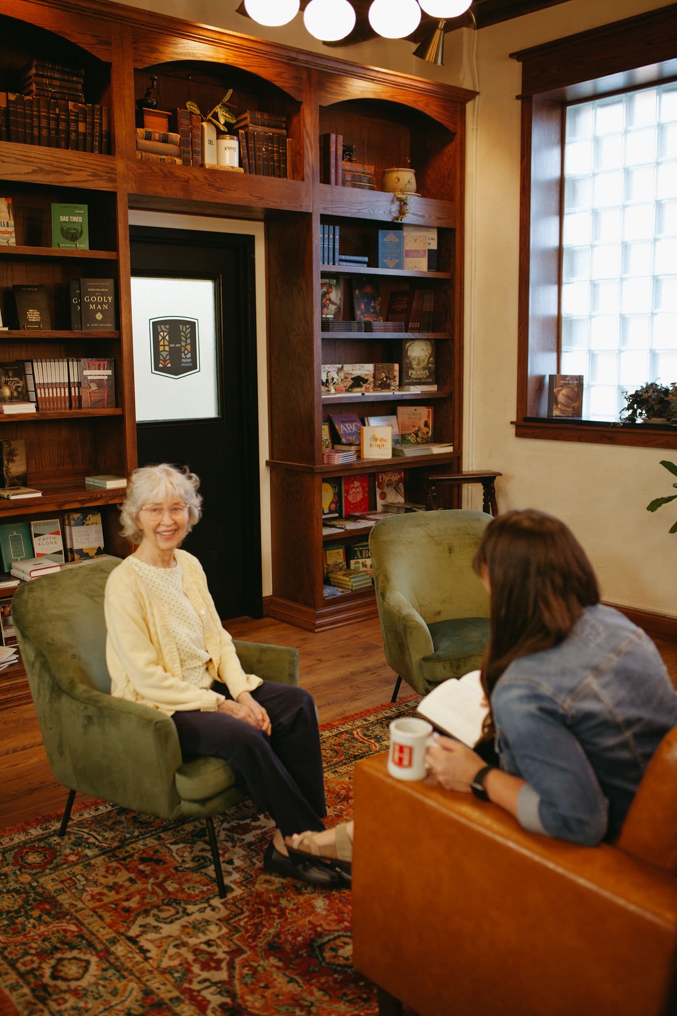 An older woman and a younger woman having a pleasant conversation in a cozy, book-lined room with comfortable chairs and a warm atmosphere.