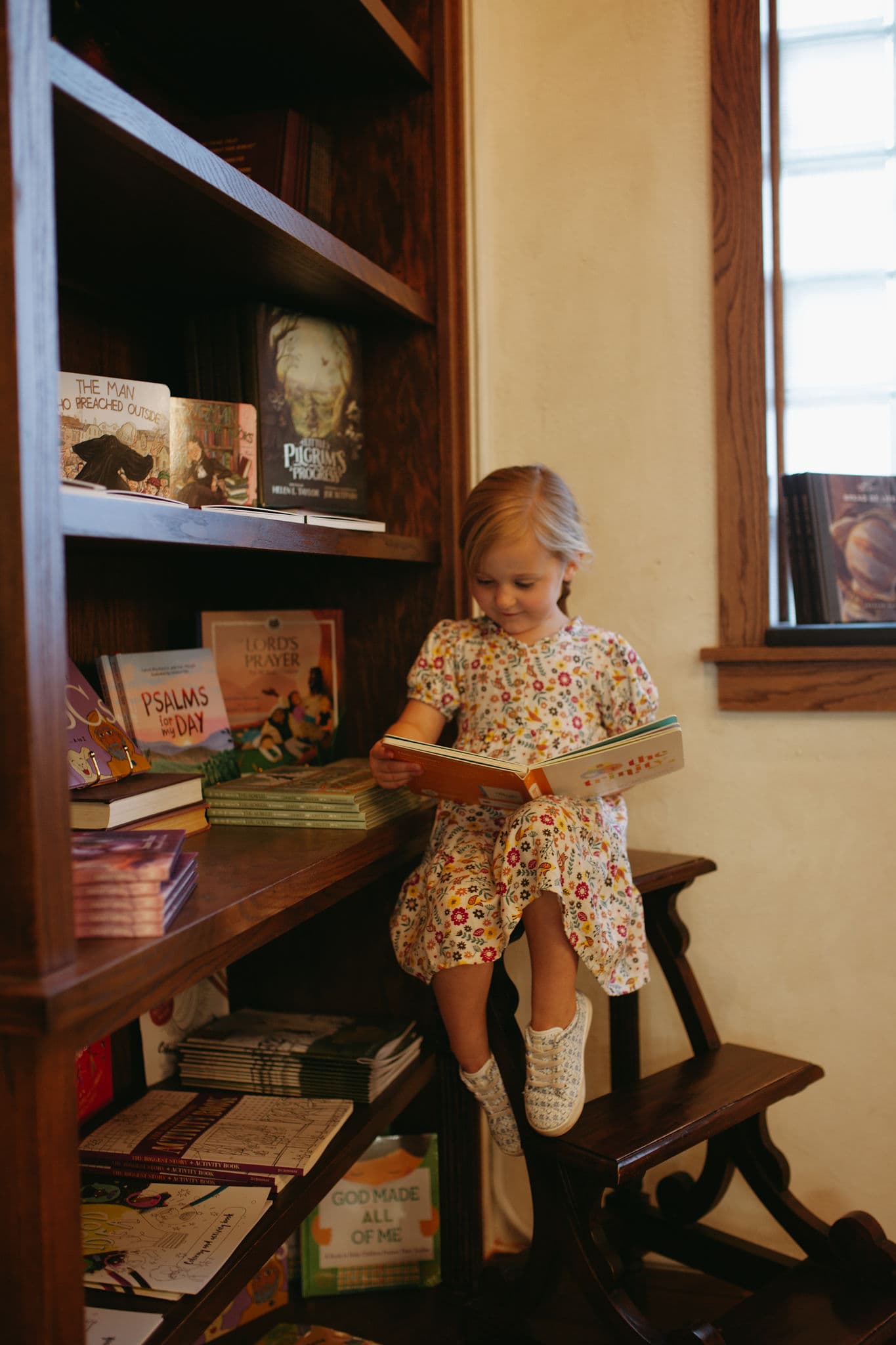 A young girl deeply engaged in reading a book while seated on a small wooden ladder in a cozy, well-lit corner of a bookstore.